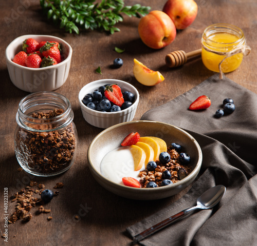 Concept of a healthy and nutritious breakfast. Delicious natural yogurt with homemade granola  peach  strawberry  honey  and blueberry in a bowl on a dark wooden background. Diet food