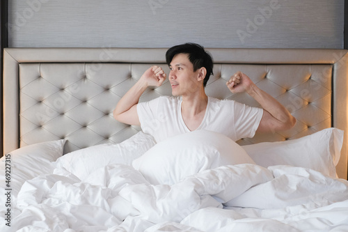 Young male sitting on bed in the morning after getting up. Handsome young man resting sitting in his bed. Young man in white t shirt feeling comfortable and sitting on bed in morning. © Avirut S.