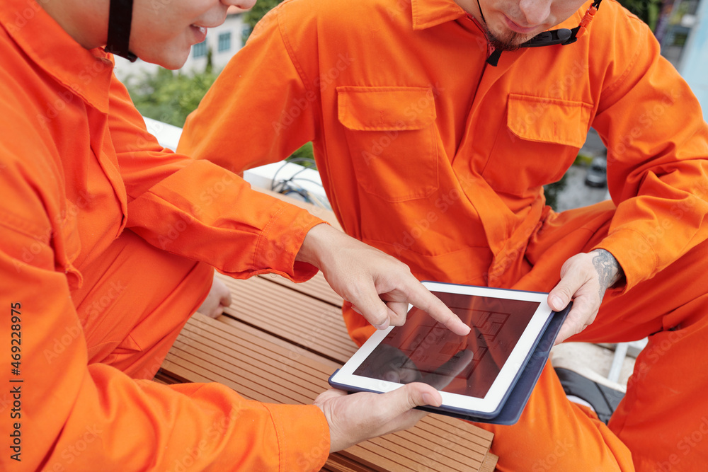 Closeup image of construction workers in bright orange overalls discussing blueprint on digital tablet