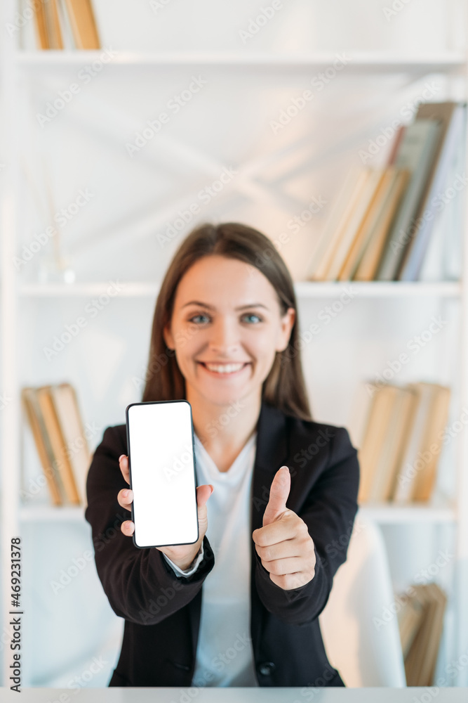 Mobile business. App advertising. Digital marketing. Satisfied smiling woman recommending white empty mockup phone screen with thumb up on defocused office background. Mobile business. App advertising. Digital marketing. Satisfied smiling woman recommending white empty mockup phone screen with thumb up on defocused office background.