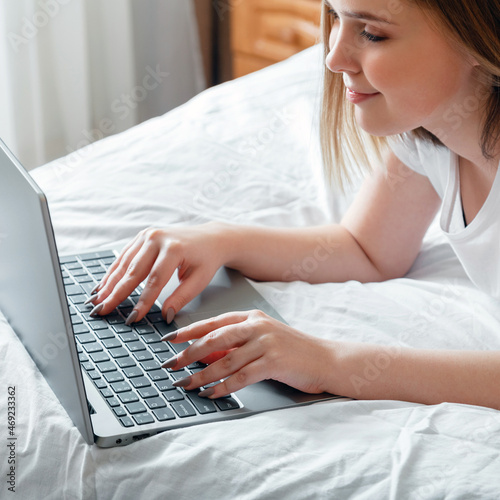 Happy teen girl communicates online working remote  studying or planning her day use computer laptop in bedroom. Young woman smile typing on laptop keyboard while lying in bed at home. Square