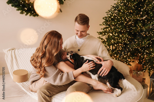 A happy couple in love and their pet dog play cuddle spend time together on Christmas holiday in the decorated interior of a room in a cozy house. Selective focus