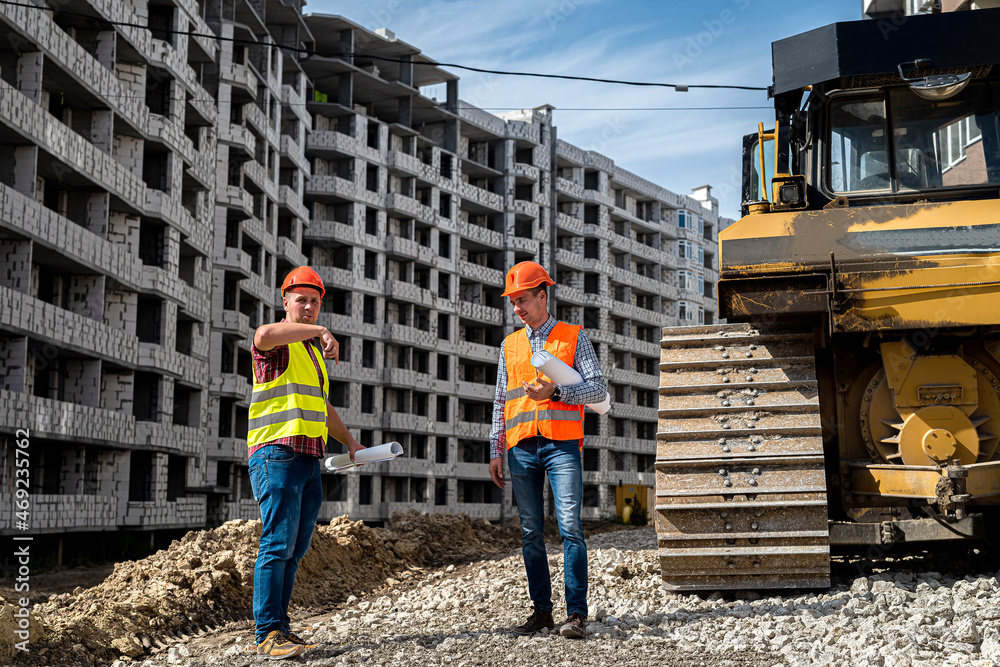 two young strong workers in uniform and helmets stand near a grader on a construction site two young strong workers in uniform and helmets stand near a grader on a construction site