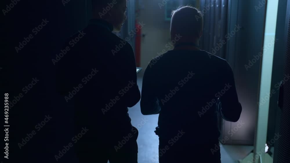 Two male engineers discussing IT project and walking in modern data office spbas. Back view of young men walk in dark server room and talk, inspect equipment or hardware and do analytical work. Multi