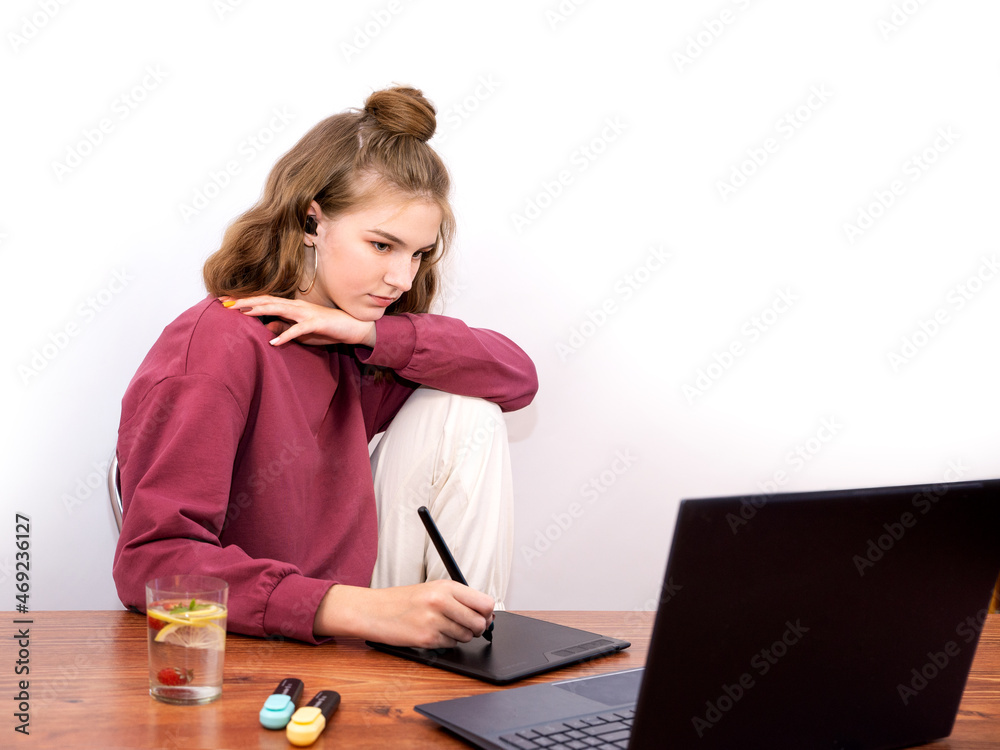 Teenage artist girl sitting at a table with a laptop draws from a graphics tablet. Hobbies and leisure of a student of a creative college. White background Teenage artist girl sitting at a table with a laptop draws from a graphics tablet. Hobbies and leisure of a student of a creative college. White background