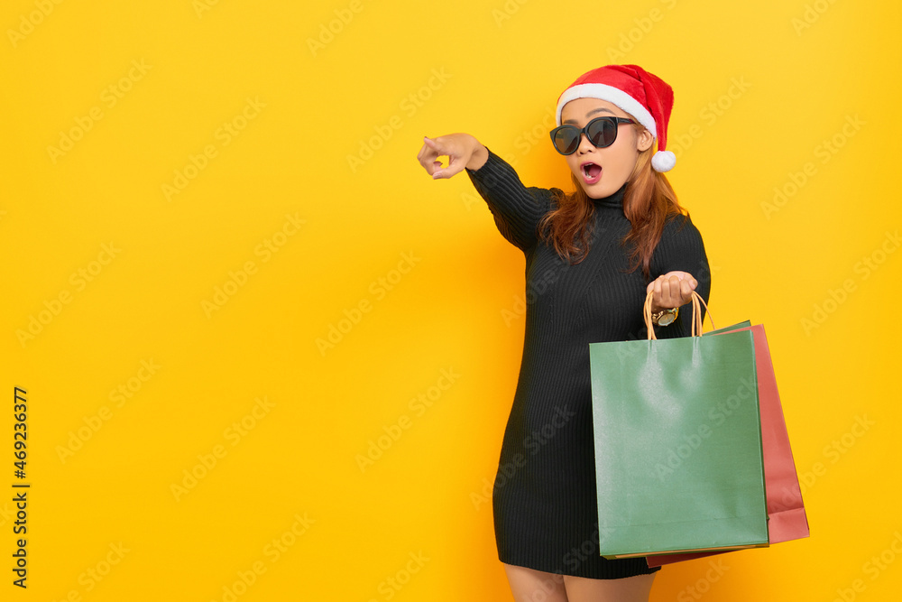 Surprised young Asian woman in Santa Claus hat and sunglasses holding shopping bags, pointing finger at copy space isolated over yellow background Surprised young Asian woman in Santa Claus hat and sunglasses holding shopping bags, pointing finger at copy space isolated over yellow background