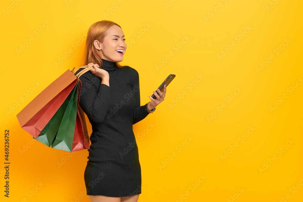 Smiling young Asian woman holding shopping bags and mobile phone looking away at copy space isolated over yellow background