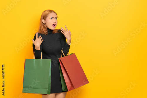 Surprised young Asian woman holding shopping bags and looking away at copy space isolated over yellow background