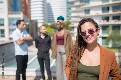 Young caucasian woman on terrace roof party. Long-haired female in casual clothes and sunglasses. People of different nationalities standing in background. Teambuilding, party concept
