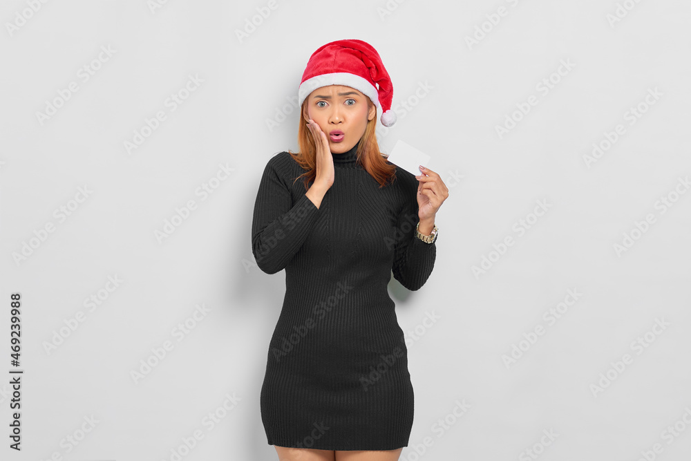 Attractive young Asian woman in Santa Claus hat showing a blank card and touching face cheek isolated over white background Attractive young Asian woman in Santa Claus hat showing a blank card and touching face cheek isolated over white background