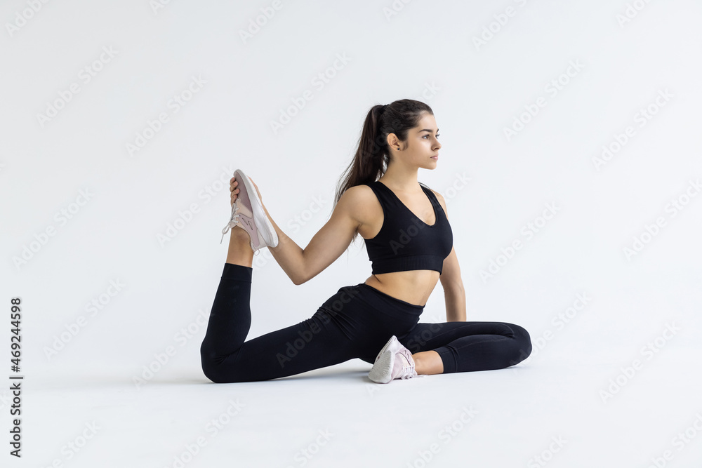 Young sporty attractive woman practicing yoga, doing Mermaid exercise, Eka Pada Rajakapotasana pose, working out, wearing sportswear, pants and top, indoor full length, white yoga studio