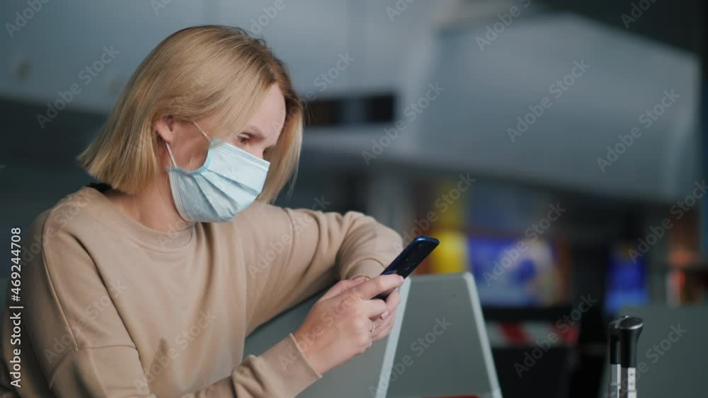 A woman in a protective mask is waiting for her flight in the airport terminal, using a smartphone A woman in a protective mask is waiting for her flight in the airport terminal, using a smartphone