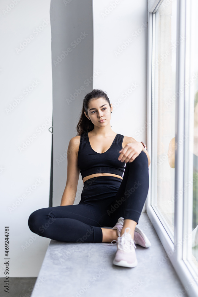 Sport, healthy lifestyle and fitness concept. Cheerful girl in sportswear, smiling with teeth, sitting windowsill, looking at window with satisfied expression Sport, healthy lifestyle and fitness concept. Cheerful girl in sportswear, smiling with teeth, sitting windowsill, looking at window with satisfied expression