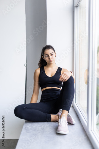 Sport, healthy lifestyle and fitness concept. Cheerful girl in sportswear, smiling with teeth, sitting windowsill, looking at window with satisfied expression