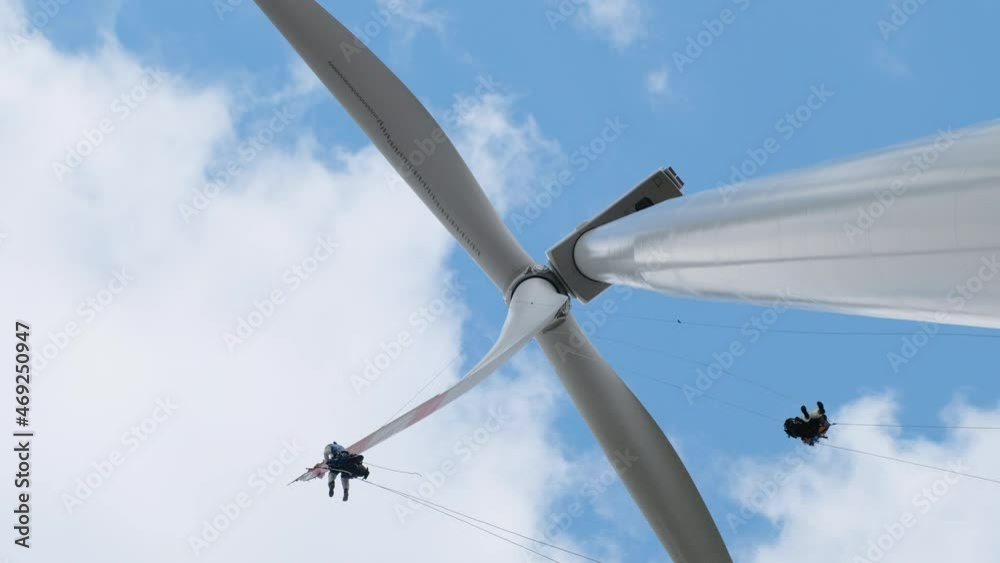 Professional high-altitude installers with climber equipment serve modern large wind generator blades under blue sky with white clouds low angle shot