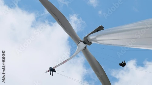 Professional high-altitude installers with climber equipment serve modern large wind generator blades under blue sky with white clouds low angle shot
