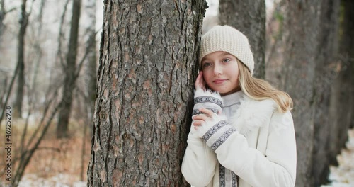 Romantic woman in white jacket and knitted hat leaned on tree trunk posing for shooting. Portrait stylish girl in winter clothes and hat posing in winter forest