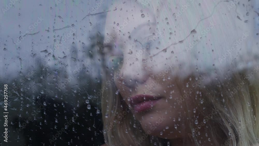 Depressed young blonde woman with mental health problems looking out of a window crying in the rain