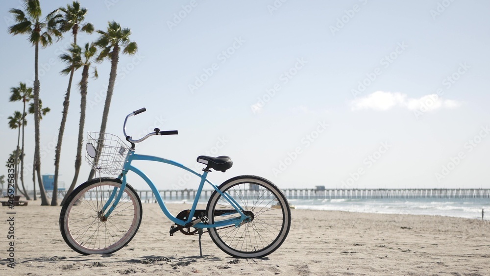 Blue bicycle, cruiser bike by sandy ocean beach, pacific coast, Oceanside pier California USA. Summertime vacations, sea shore. Vintage cycle, palms, sky, lifeguard tower watchtower hut, car truck.