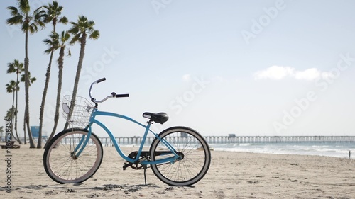 Blue bicycle, cruiser bike by sandy ocean beach, pacific coast, Oceanside pier California USA. Summertime vacations, sea shore. Vintage cycle, palms, sky, lifeguard tower watchtower hut, car truck. photo