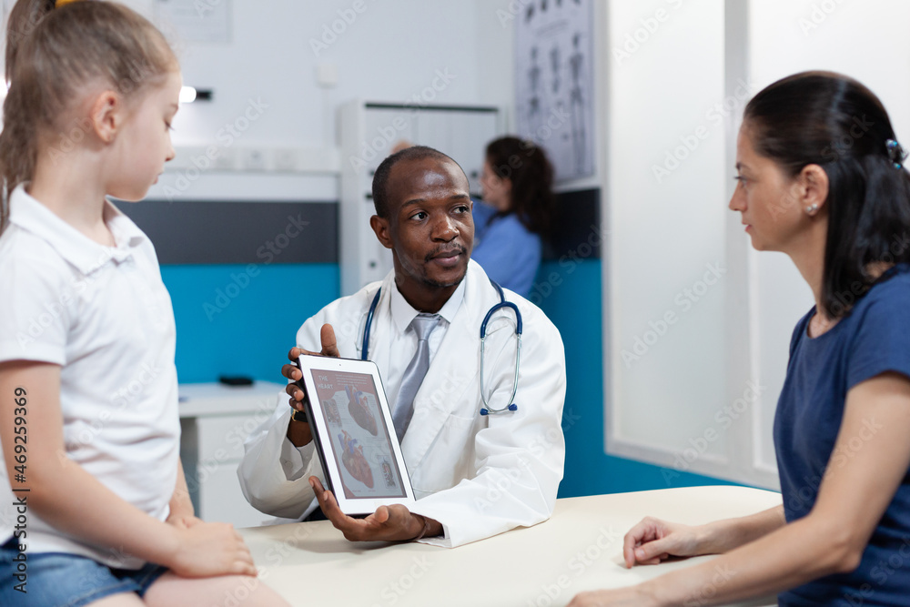 African american radiologist doctor holding tablet computer with heart radiography on screen discussing xray expertise results with mother during examination.Therapist man working in hospital office African american radiologist doctor holding tablet computer with heart radiography on screen discussing xray expertise results with mother during examination.Therapist man working in hospital office