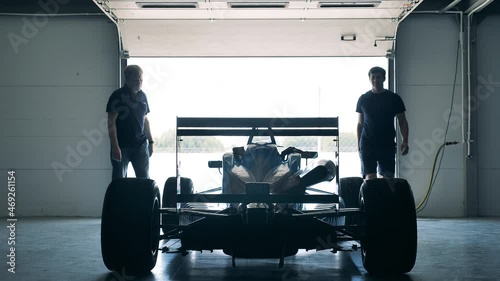 Two men open the garage and wheel a racing car out of it