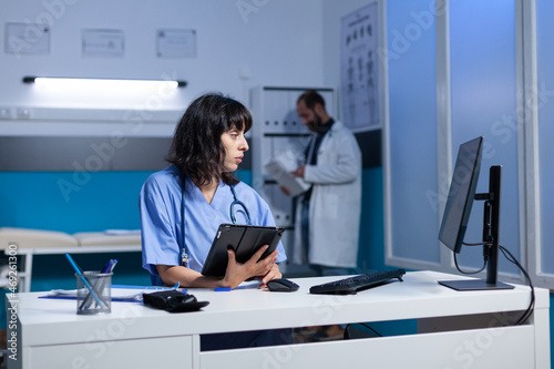 Woman working as nurse with tablet for checkup at night. Medical assistant looking at modern device and computer on desk for healthcare and treatment, working late. Person with job
