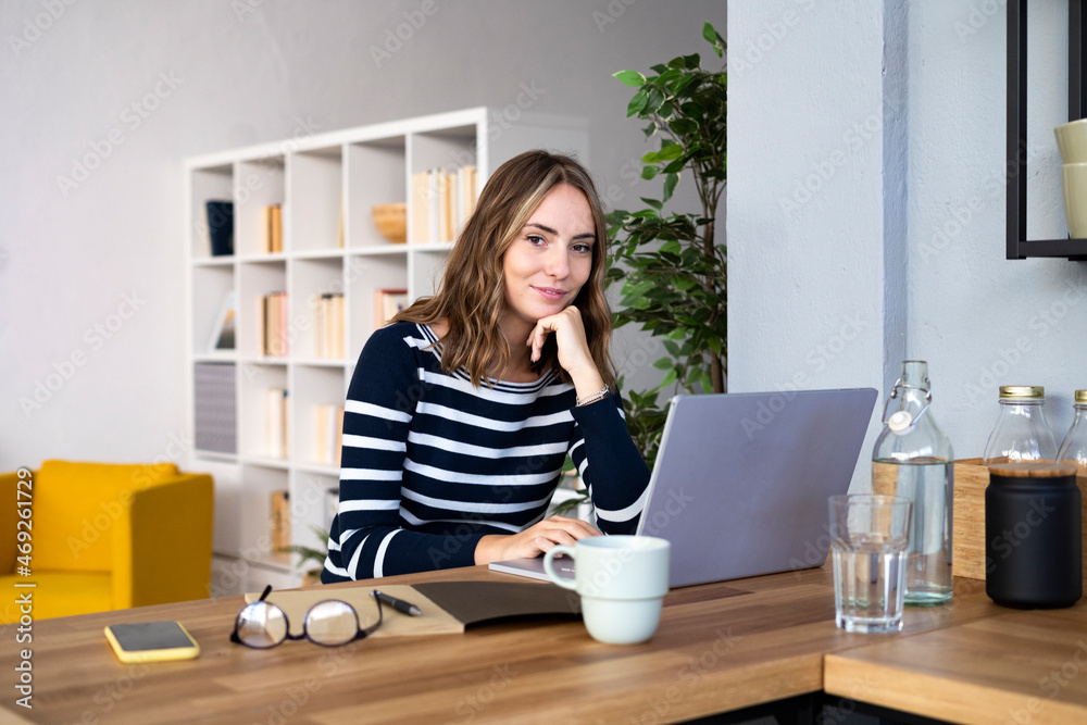 Young woman with hand on chin in front of laptop at kitchen Young woman with hand on chin in front of laptop at kitchen