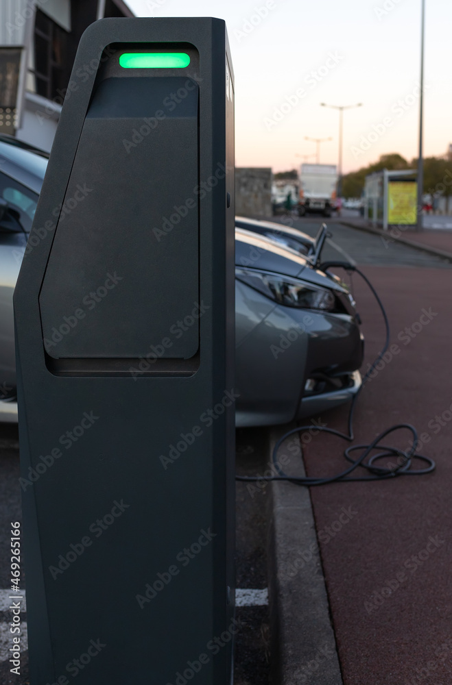 Close-up of a charging station for electric cars in a public street car park in a French city. Behind the charging station is a grey electric car charging, out of focus. Close-up of a charging station for electric cars in a public street car park in a French city. Behind the charging station is a grey electric car charging, out of focus.