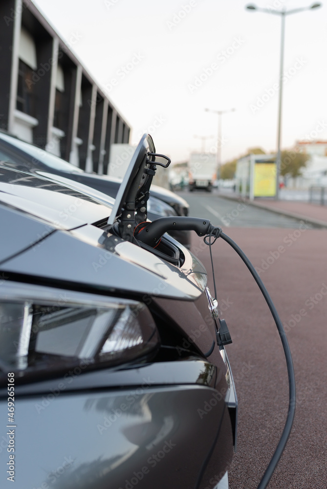 Grey electric car charging from the front on a street in a French city. Charging port door open with charging plug connected. Close-up with blurred background.