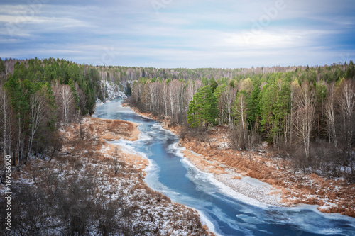 Picturesque harsh winter or late autumn nature landscape with blue river going through pine forest with banks covered with dried yellow grass at cloudy frosty day with heavy sky. Horizontal image