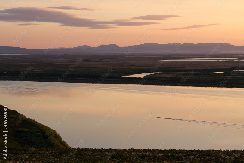 Evening view of the river valley. Summer arctic landscape. A boat is sailing along a wide river. In the distance tundra and mountains. The nature of Chukotka and Siberia. Anadyr River, Far East Russia