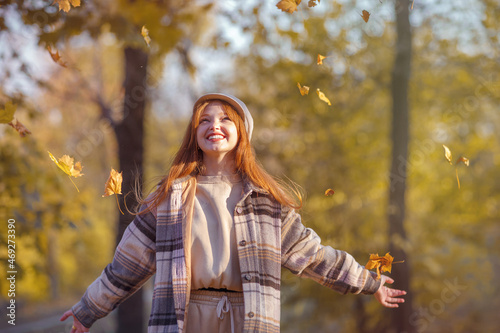 A young pretty girl with long red hair walks in the park,throws yellow leaves into the air and laughs happily, expressing joy , happiness. Joy, autumn, sunny day, vacation concept.