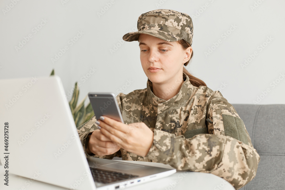 Horizontal shot of calm woman soldier wearing camouflage uniform and hat, posing at home, sitting in front of portable computer and using mobile phone, has calm expression. Horizontal shot of calm woman soldier wearing camouflage uniform and hat, posing at home, sitting in front of portable computer and using mobile phone, has calm expression.
