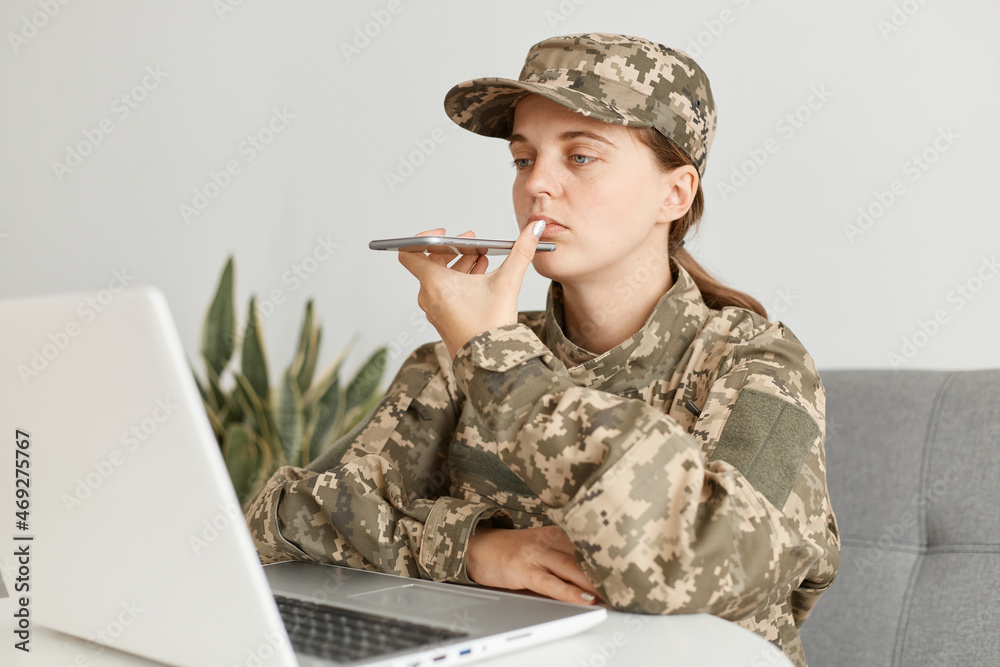 Portrait of Caucasian female soldier wearing military costume, posing indoor in light room, recording audio message, using voice assistant for writing command.