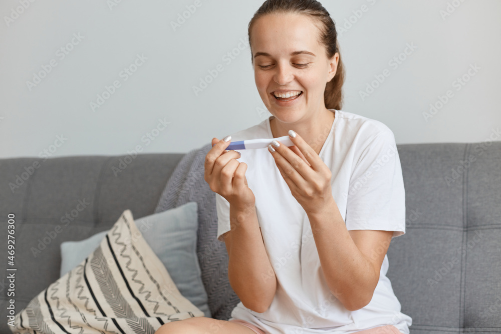 Portrait of smiling Caucasian dark haired female wearing casual style clothing with pregnancy test in hands, rejoicing positive result, posing at home while sitting in living room.