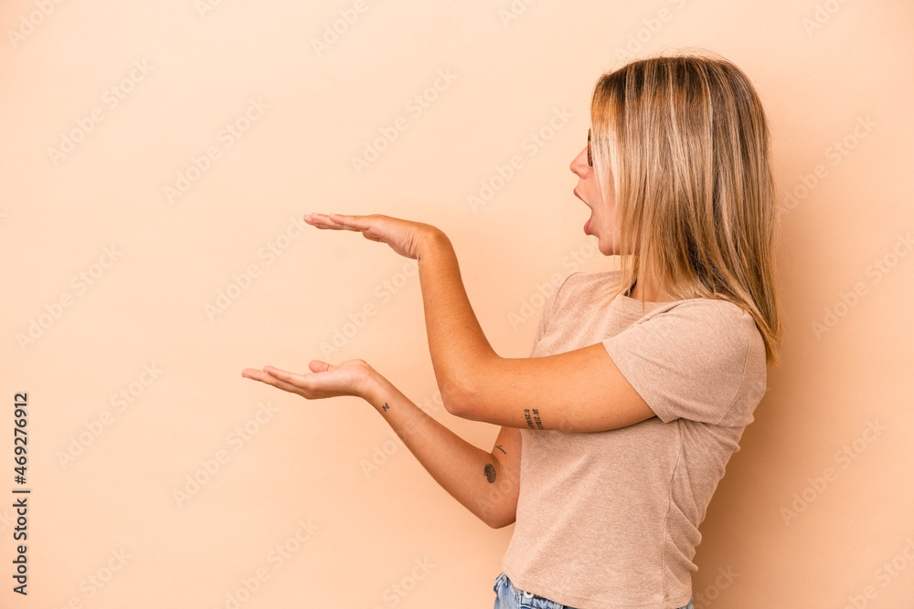 Young caucasian woman isolated on beige background shocked and amazed holding a copy space between hands. Young caucasian woman isolated on beige background shocked and amazed holding a copy space between hands.