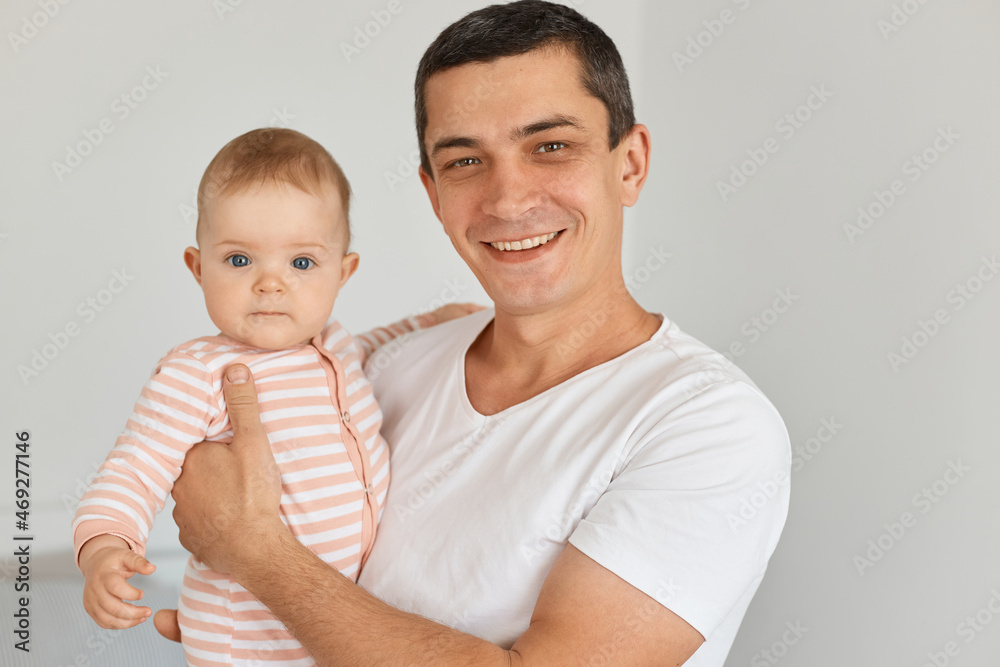 Portrait of smiling happy brunette man wearing white casual t shirt holding his cute infant daughter, posing at home in light room, looking at camera, expressing positive emotions. Portrait of smiling happy brunette man wearing white casual t shirt holding his cute infant daughter, posing at home in light room, looking at camera, expressing positive emotions.