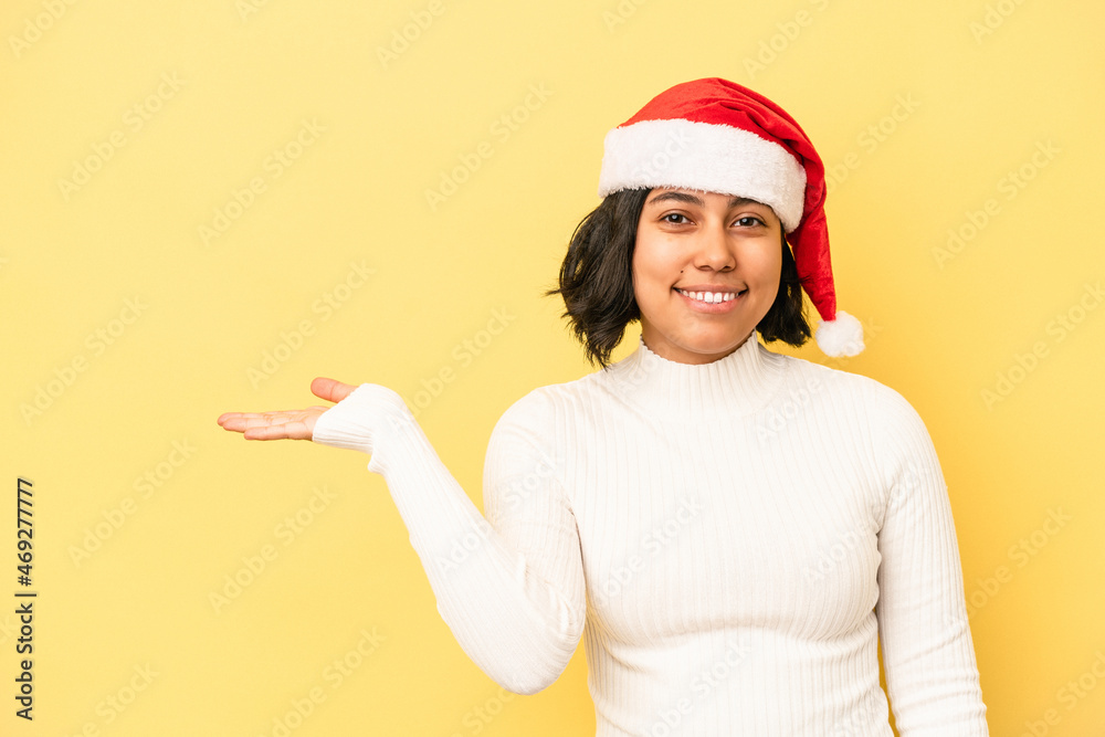 Young latin woman celebrating christmas isolated on yellow background showing a copy space on a palm and holding another hand on waist. Young latin woman celebrating christmas isolated on yellow background showing a copy space on a palm and holding another hand on waist.