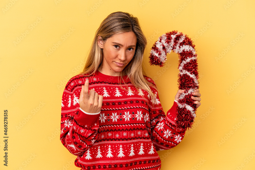 Young caucasian woman holding big christmas stick isolated on yellow background pointing with finger at you as if inviting come closer. Young caucasian woman holding big christmas stick isolated on yellow background pointing with finger at you as if inviting come closer.