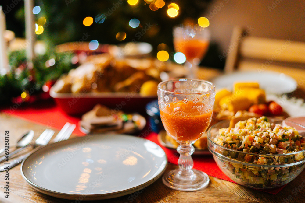 Close-up of glass with fresh orange juice and delicious vegetable salad on feast Christmas dinner table during holiday friendly family party, on blurred background of xmas celebration bokeh lights.