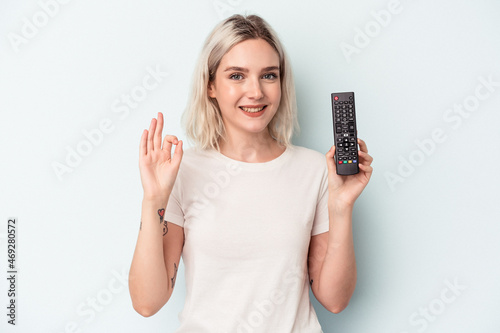 Young caucasian woman holding a tv controller isolated on blue background cheerful and confident showing ok gesture.