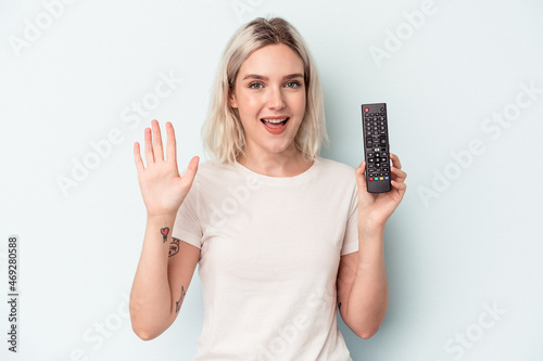 Young caucasian woman holding a tv controller isolated on blue background smiling cheerful showing number five with fingers.