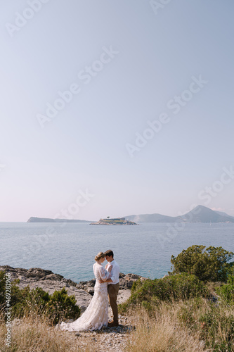 Groom and bride stand embracing on the seashore against the backdrop of the mountains