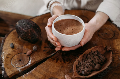Hot handmade ceremonial cacao in white cup. Woman hands holding craft cocoa, top view on wooden table. Organic healthy chocolate drink prepared from beans, no sugar. Giving cup on ceremony, cozy cafe photo
