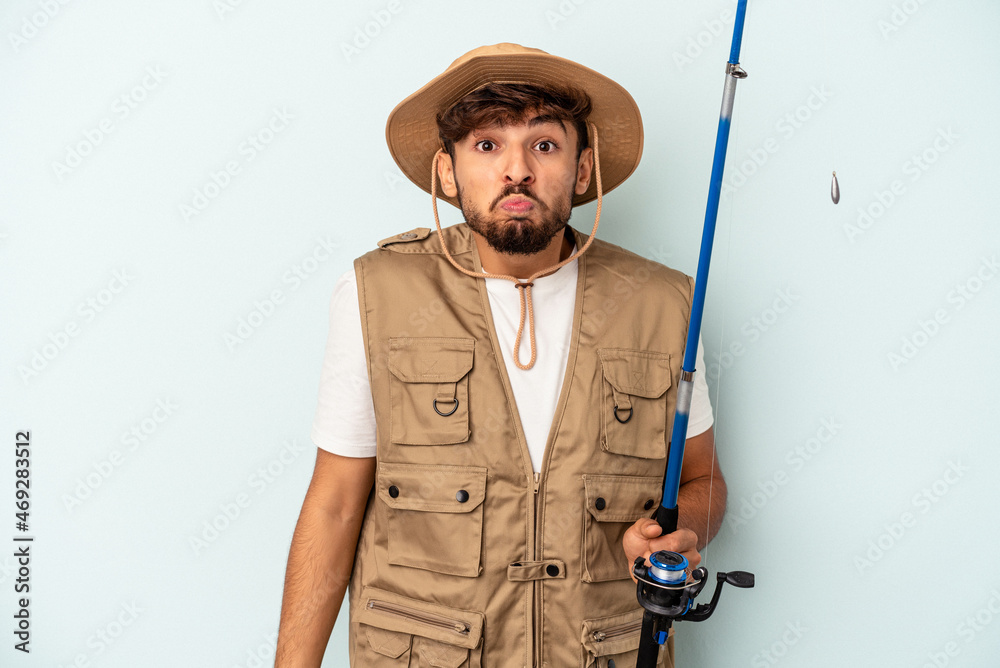 Young mixed race fisherman holding a rod isolated on blue background shrugs shoulders and open eyes confused. Young mixed race fisherman holding a rod isolated on blue background shrugs shoulders and open eyes confused.