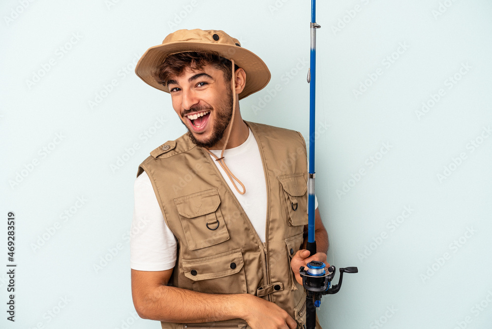 Young mixed race fisherman holding a rod isolated on blue background laughing and having fun. Young mixed race fisherman holding a rod isolated on blue background laughing and having fun.
