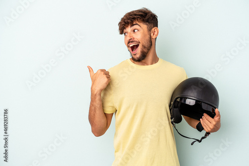 Young mixed race man holding motorcycle helmet isolated on blue background points with thumb finger away, laughing and carefree.