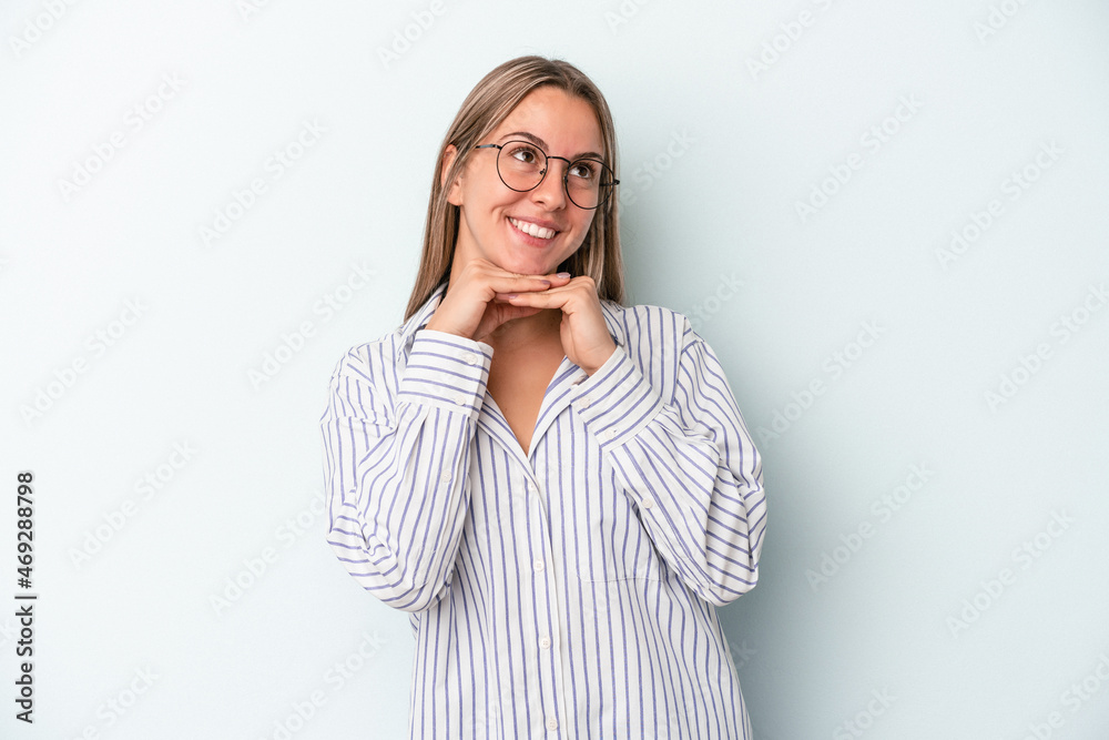 Young caucasian woman isolated on blue background keeps hands under chin, is looking happily aside. Young caucasian woman isolated on blue background keeps hands under chin, is looking happily aside.