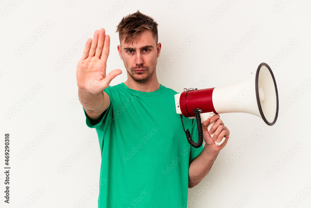 Young caucasian man holding megaphone isolated on white background standing with outstretched hand showing stop sign, preventing you. Young caucasian man holding megaphone isolated on white background standing with outstretched hand showing stop sign, preventing you.